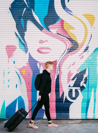 boy in black dragging a suitcase behind him walking in front of a wall covered with graffiti